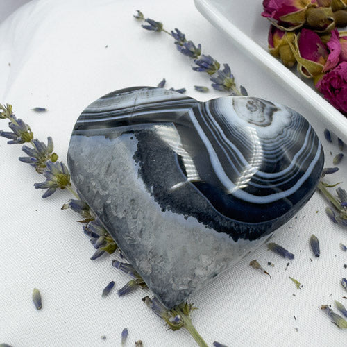 Heart shaped black stripe agate stone surrounded by dried lavender and rose petals on white surface