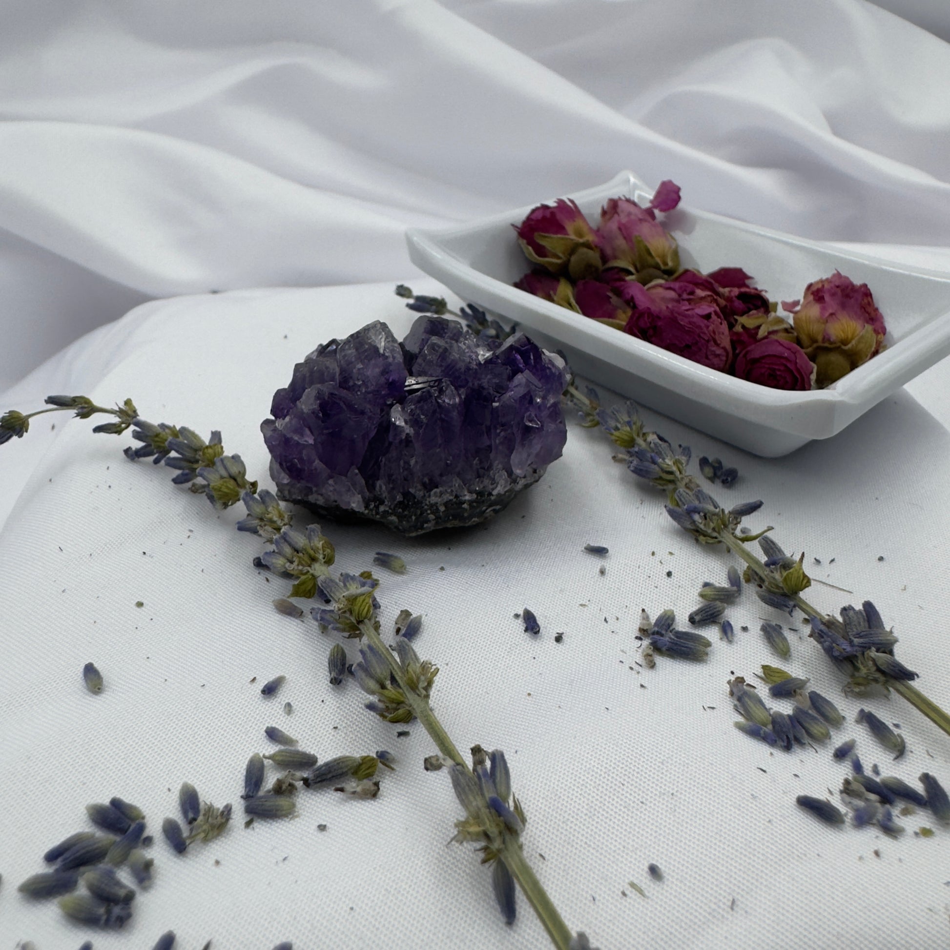 Davinas amethyst raw crystal cluster on white fabric with dried lavender and rose petals nearby