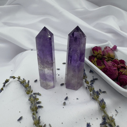 Two polished purple Amethyst Tower Point crystals standing on white fabric with dried flowers nearby