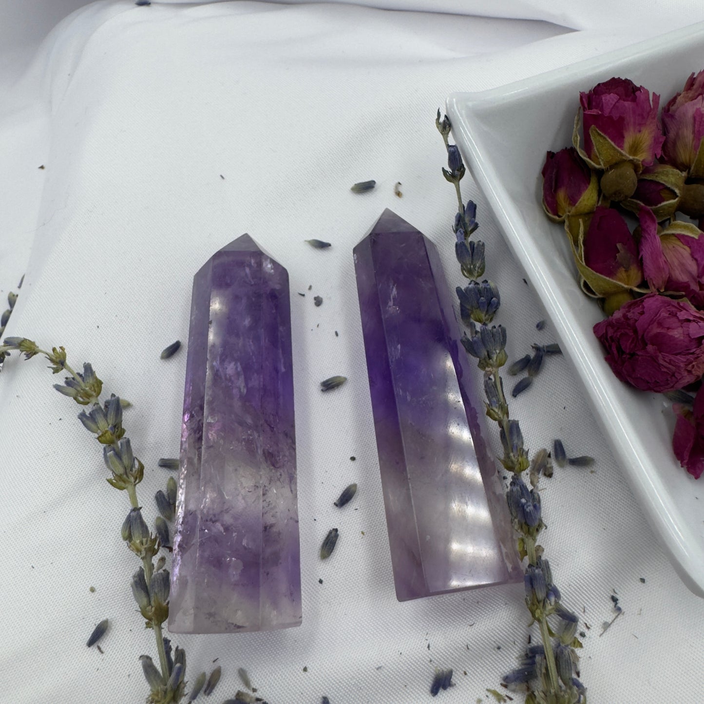 Two polished Amethyst Tower Point crystals placed on white fabric with dried lavender and rose buds