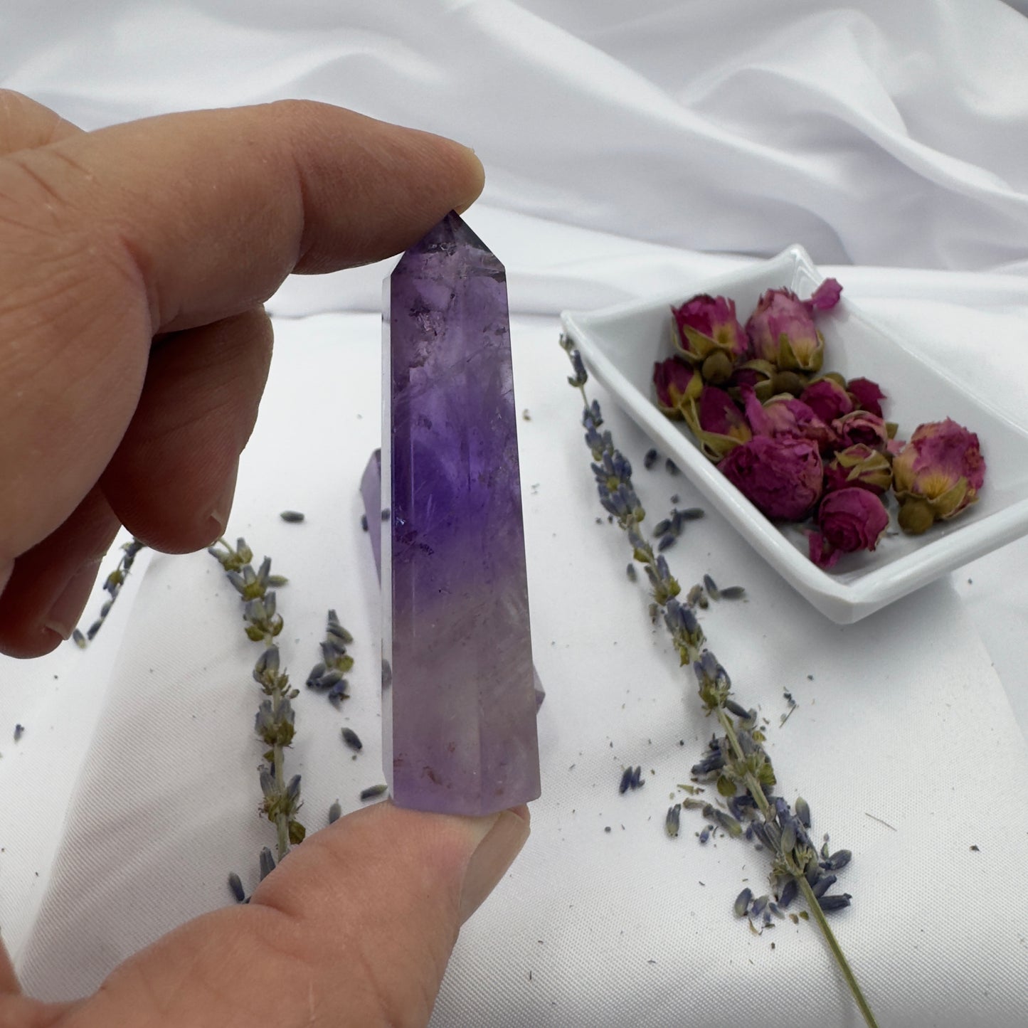 Davinas Amethyst Tower Point held between fingers with dried flowers and roses on white fabric background
