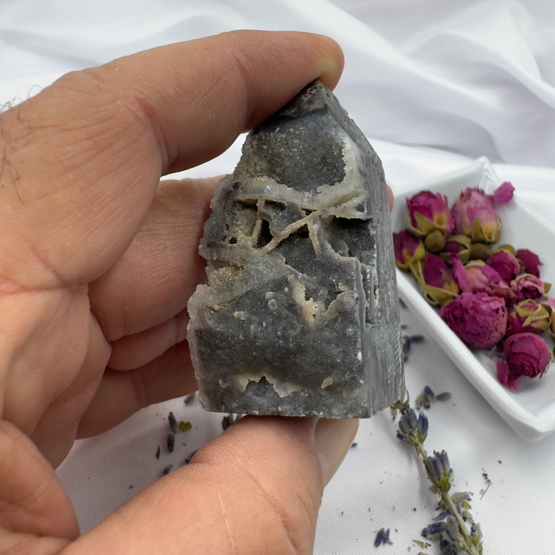 Hand holding a Black Sphalerite Geode Tower with natural crystal formations and dried flowers in background