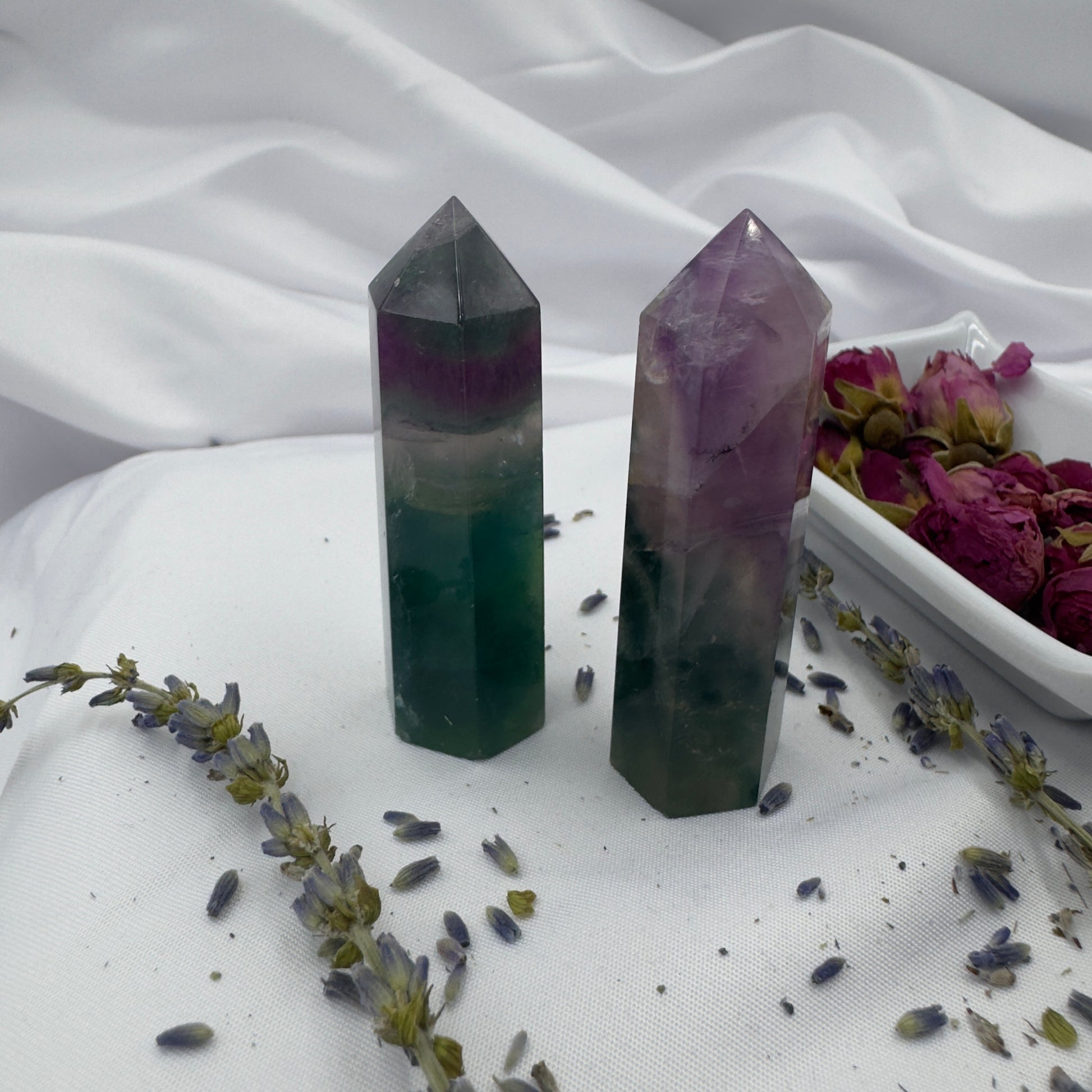 Two polished blue purple green fluorite point crystals standing on white cloth beside dried flowers