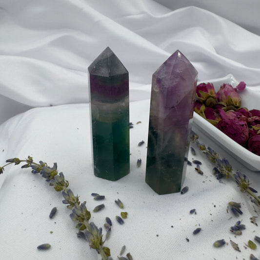 Two polished blue purple green fluorite point crystals standing on white cloth beside dried flowers