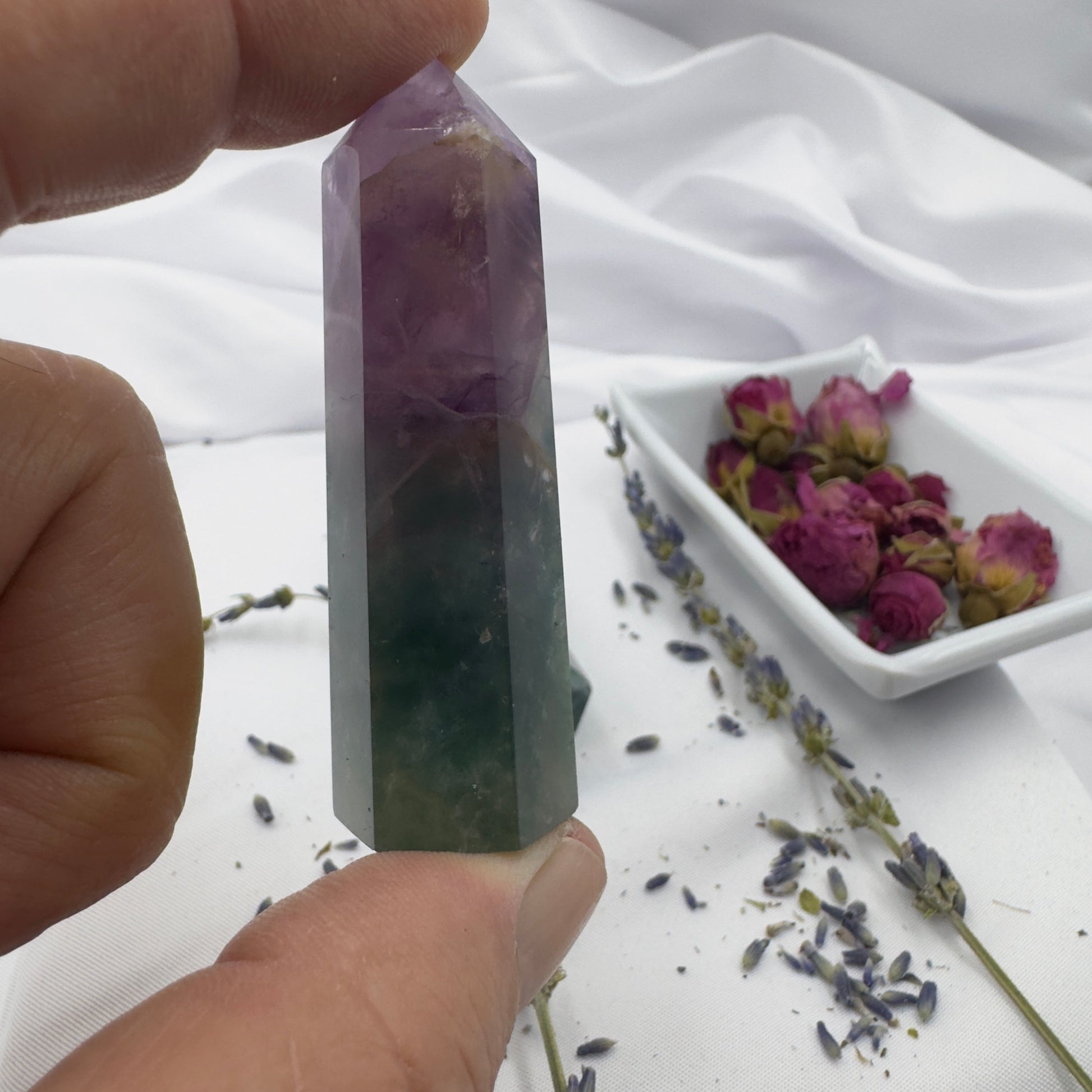 Davinas blue purple green fluorite point held between fingers with dried flowers on white fabric background