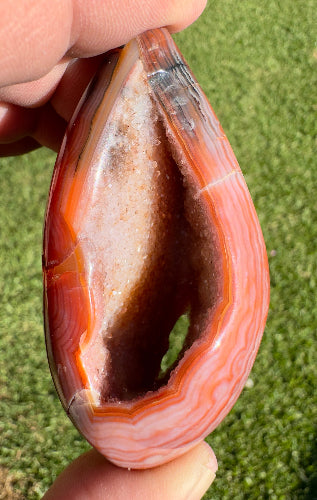 Davinas Carnelian Geode Druzy Freeform gemstone with vibrant orange and white layers held in hand against grass background