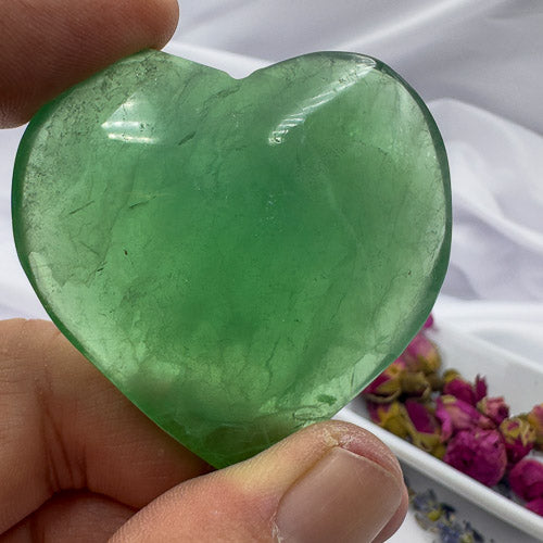 Hand holding a polished green fluorite crystal heart with visible natural patterns and a soft white background