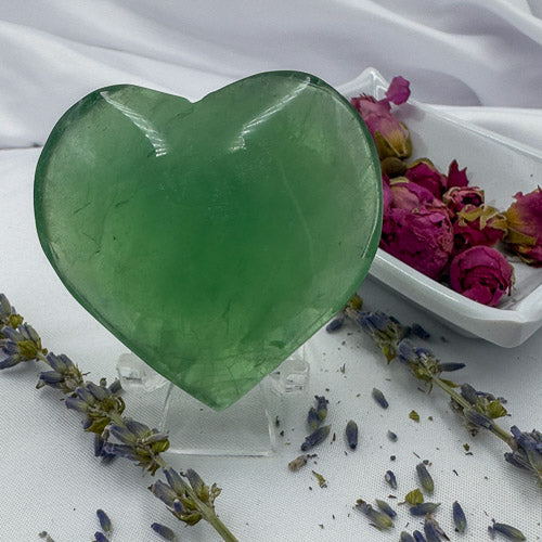 Green fluorite crystal heart displayed on a clear stand with dried lavender and rose petals nearby