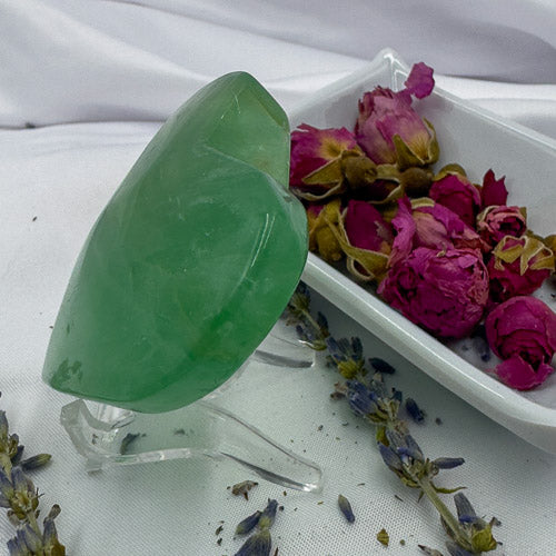 Davinas heart shaped fluorite crystal in green displayed on a clear stand with dried flowers in the background