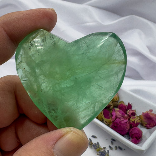 Close-up of a hand holding a heart shaped fluorite crystal with a white cloth and dried flowers in the background
