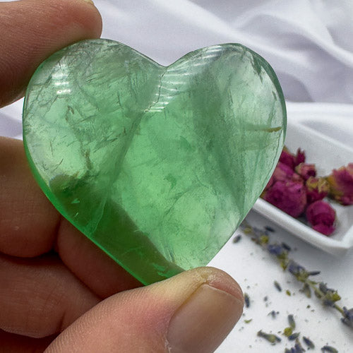 Hand holding a polished heart shaped green fluorite stone with dried flowers in the background