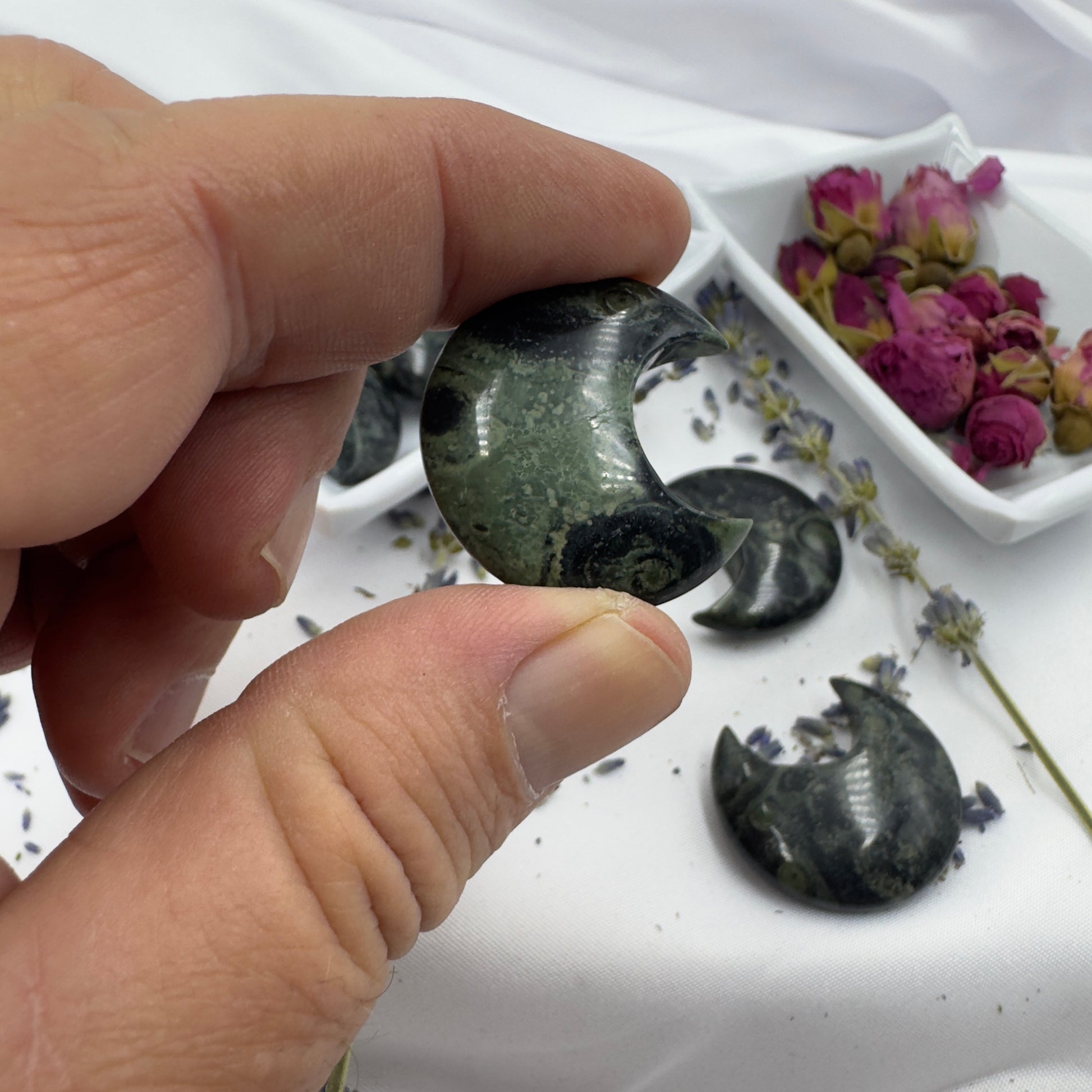 Hand holding a crescent-shaped Kambaba Jasper Moon stone with dried flowers in background