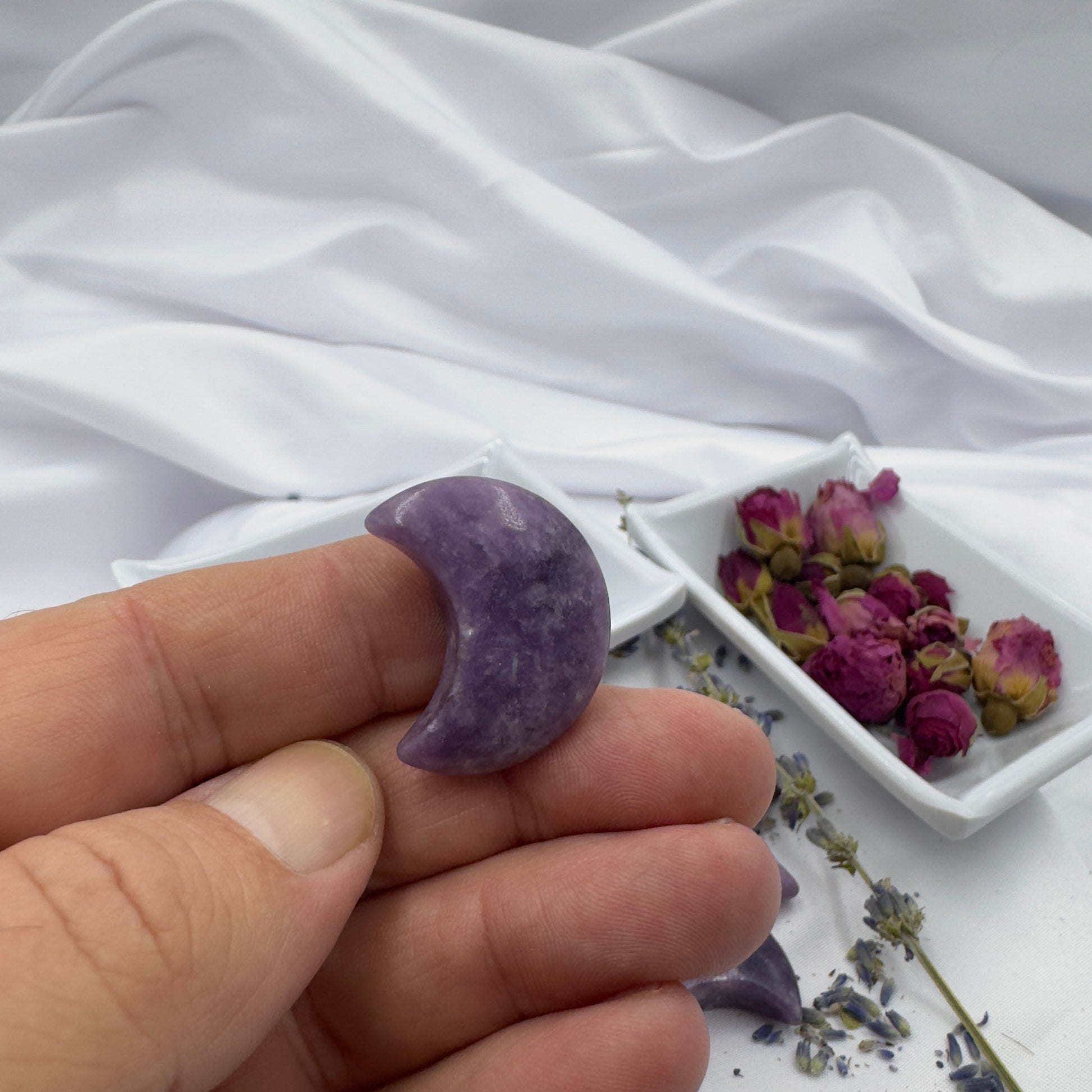 Hand holding a small polished Lepidolite moon crystal shaped like a crescent moon with dried flowers in the background