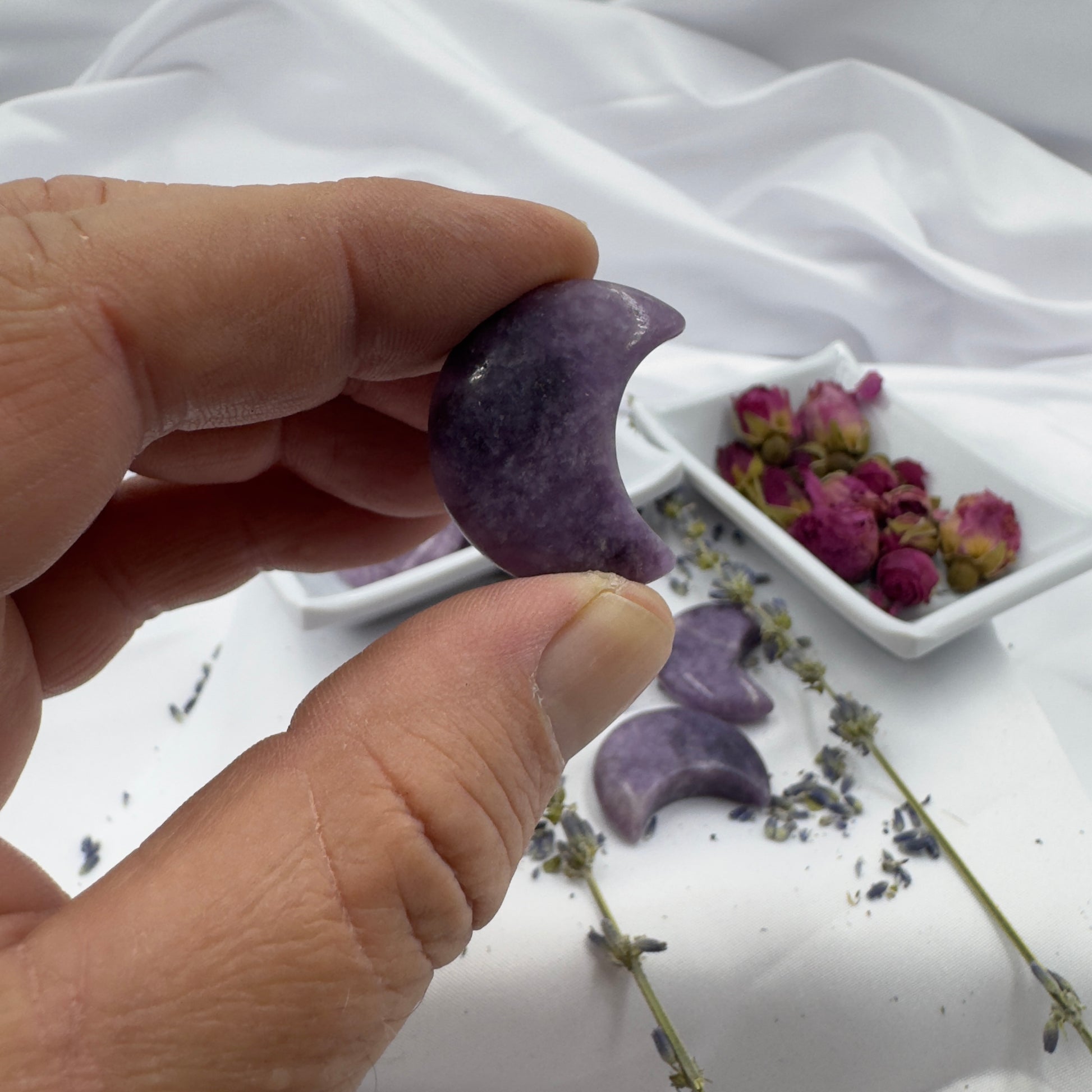 Hand holding a polished Lepidolite moon crystal carved in a crescent shape with dried flowers in the background