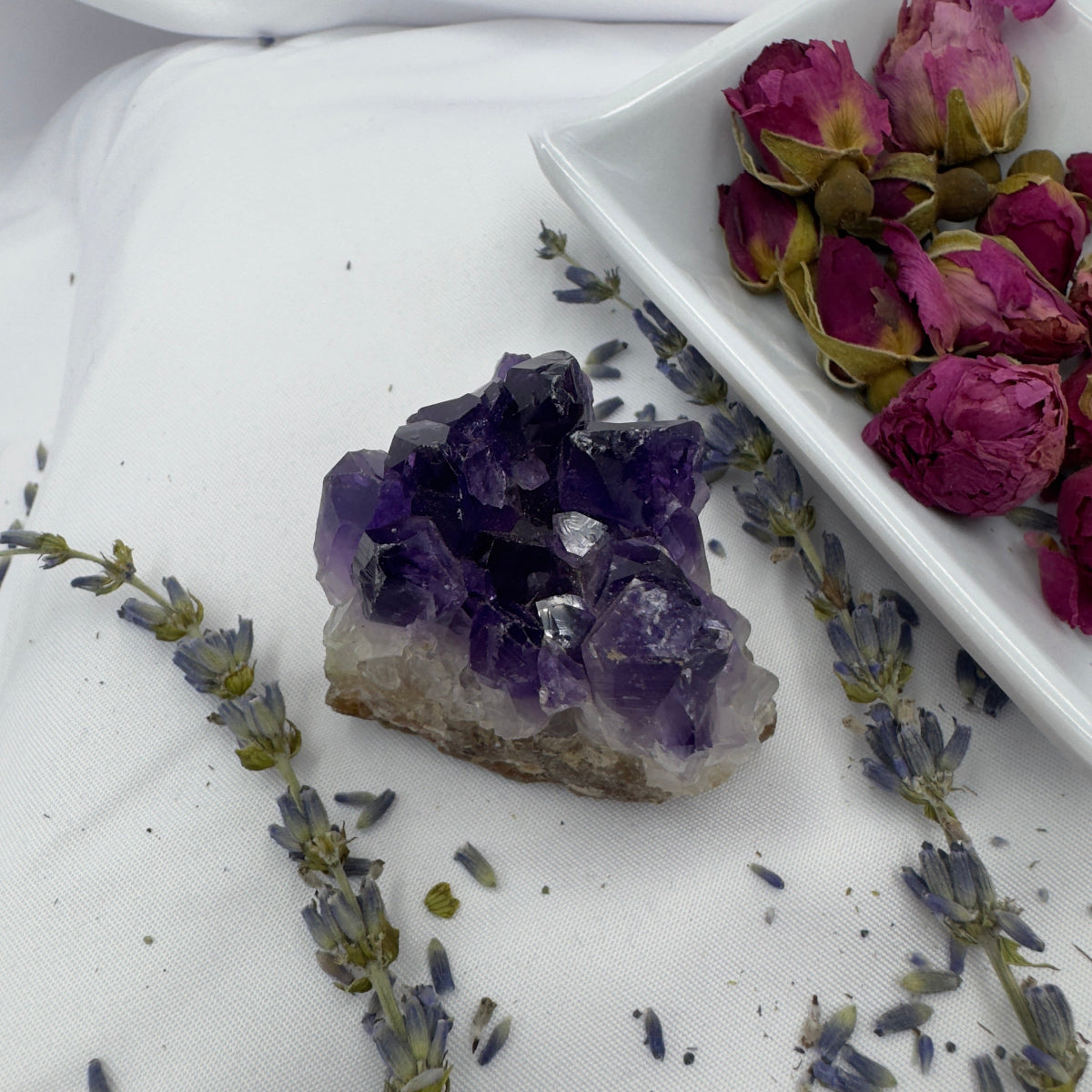 Davinas amethyst raw crystal cluster surrounded by dried lavender and rose buds on a white surface