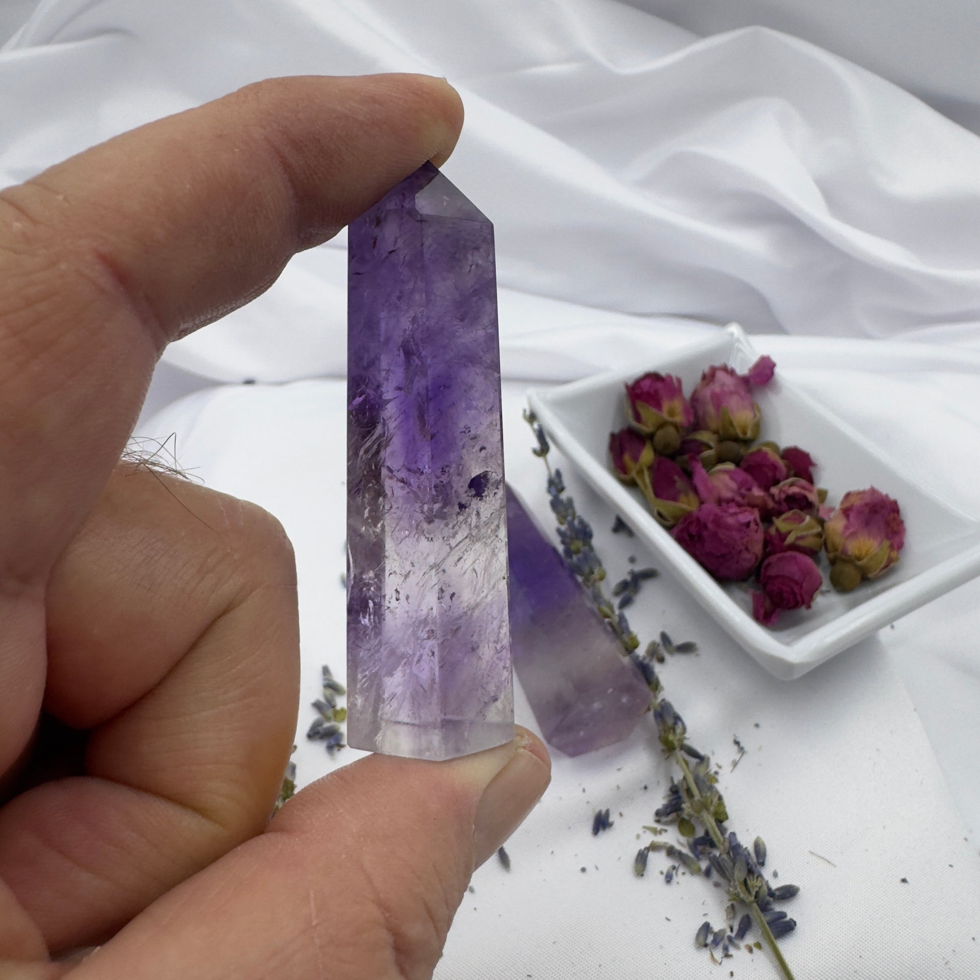Hand holding a polished Amethyst Tower Point crystal with dried rosebuds and lavender on white fabric background
